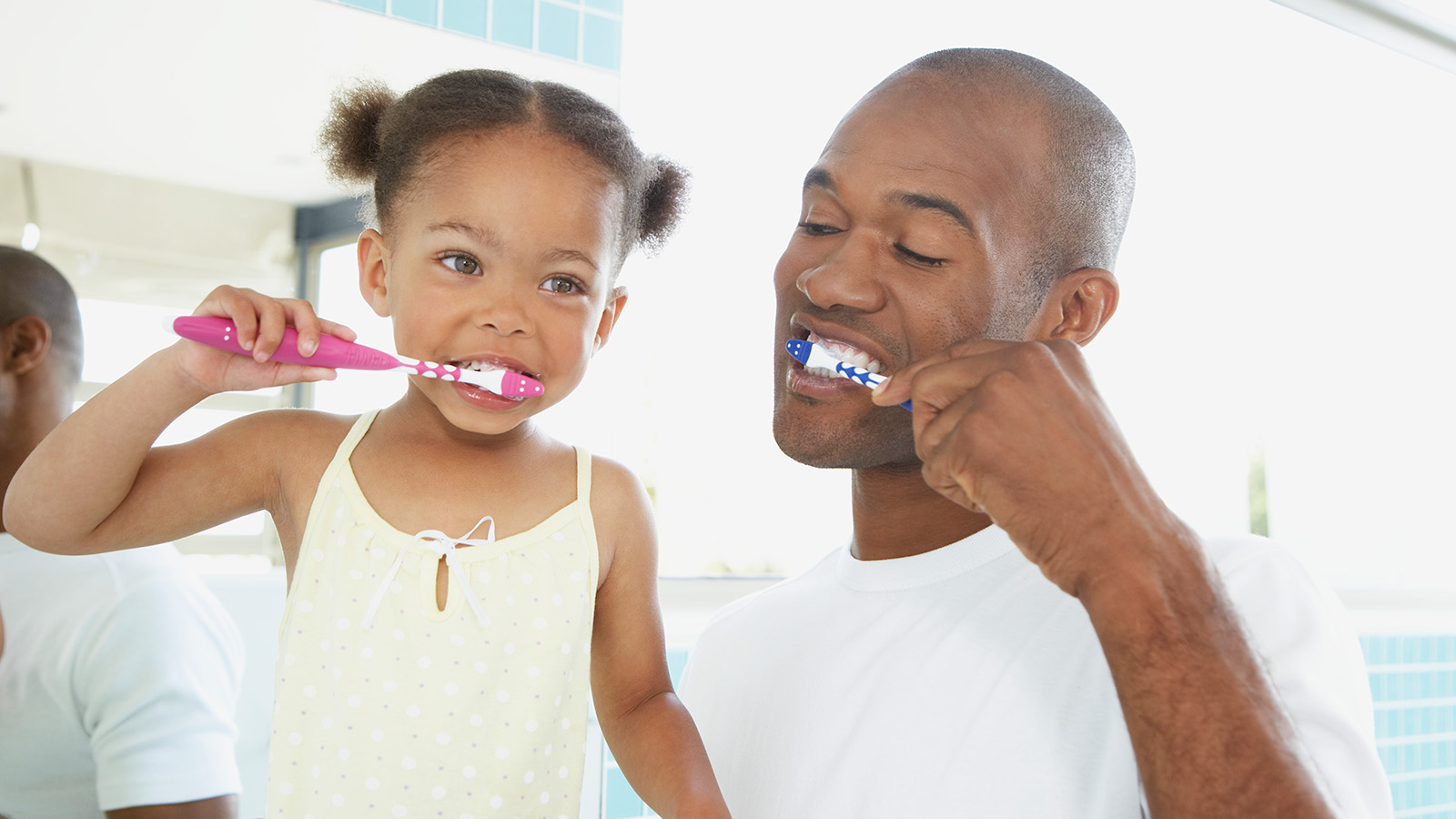 A father and daughter brush their teeth together in a bright bathroom, smiling at each other.