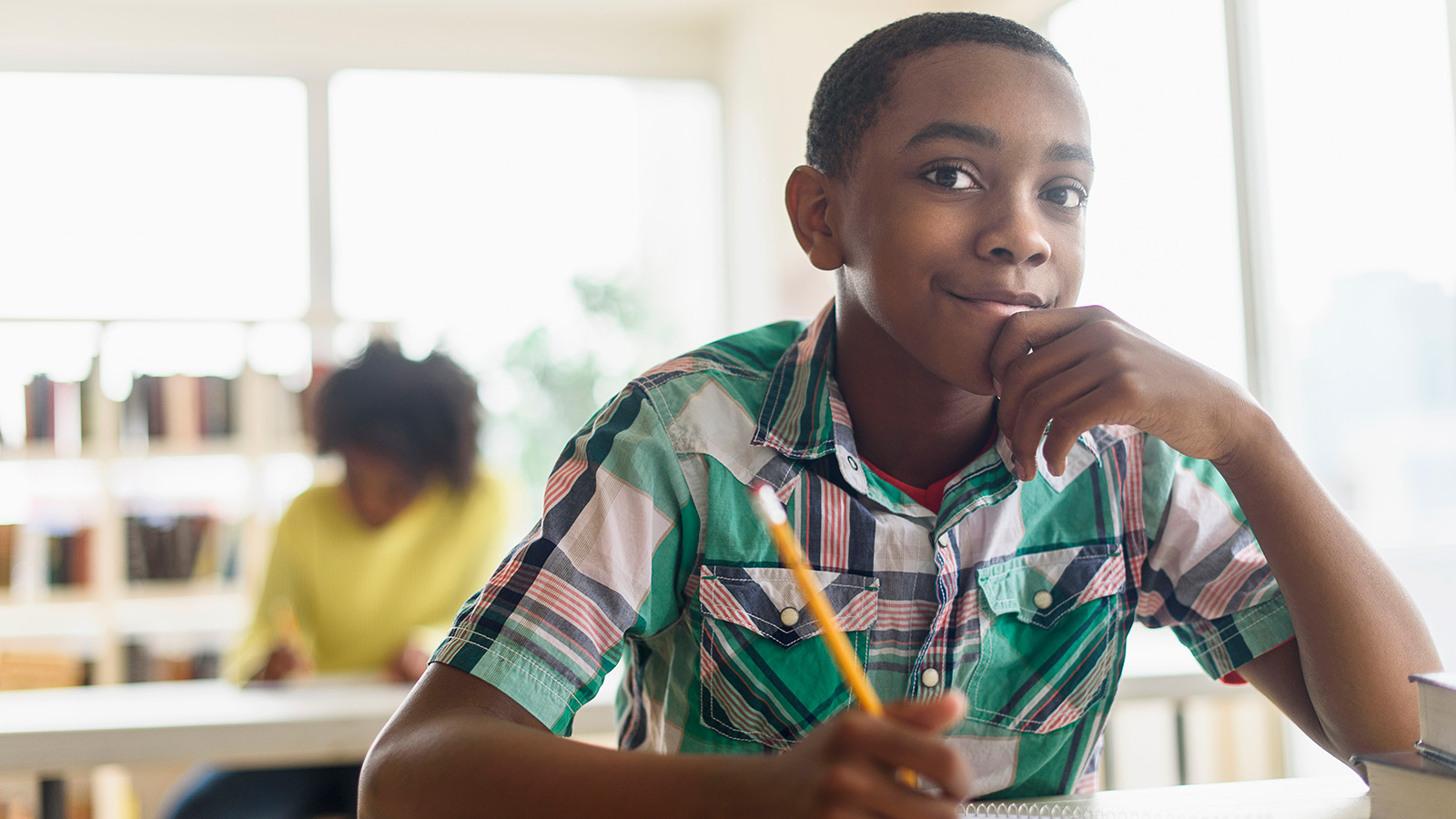A child in a colorful shirt sits at a desk, holding a pencil, focused on their notebook in a classroom.