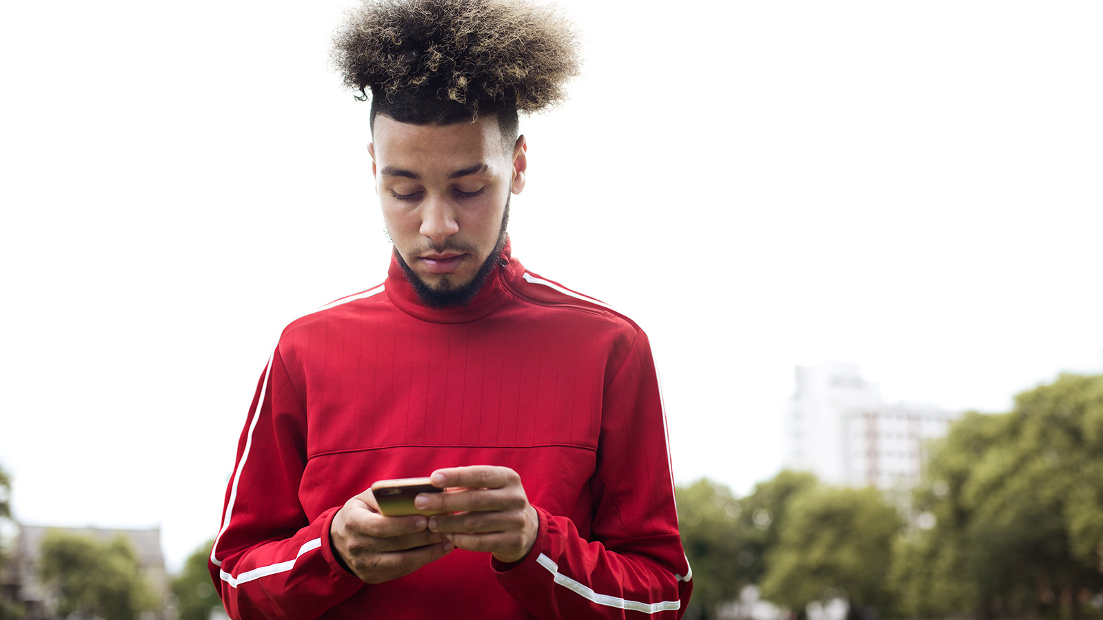 A young man stands outdoors, wearing a red athletic shirt, looking at his phone with a city backdrop behind him.