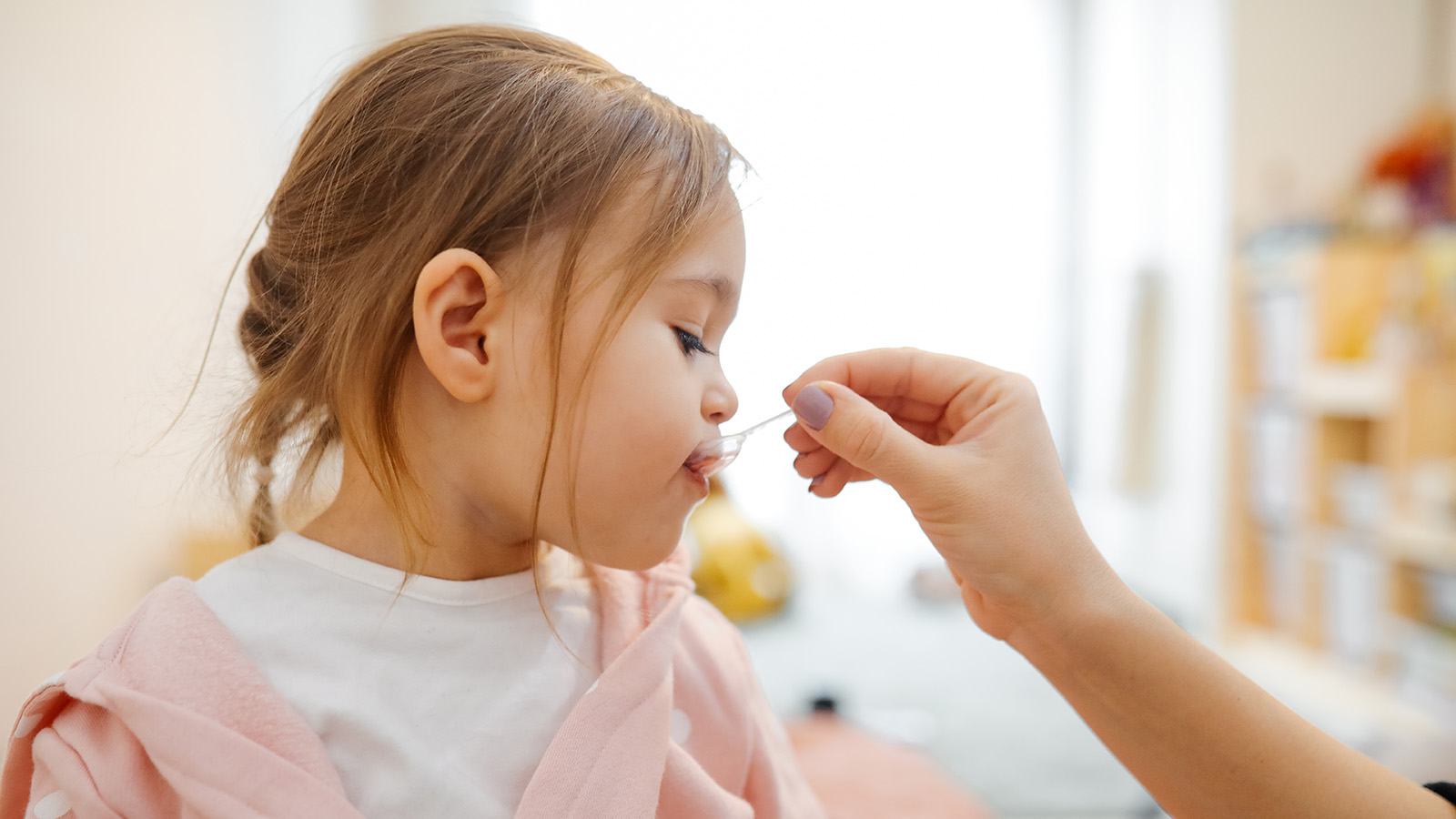 A woman feeds liquid medicine to a young girl in a bright room.