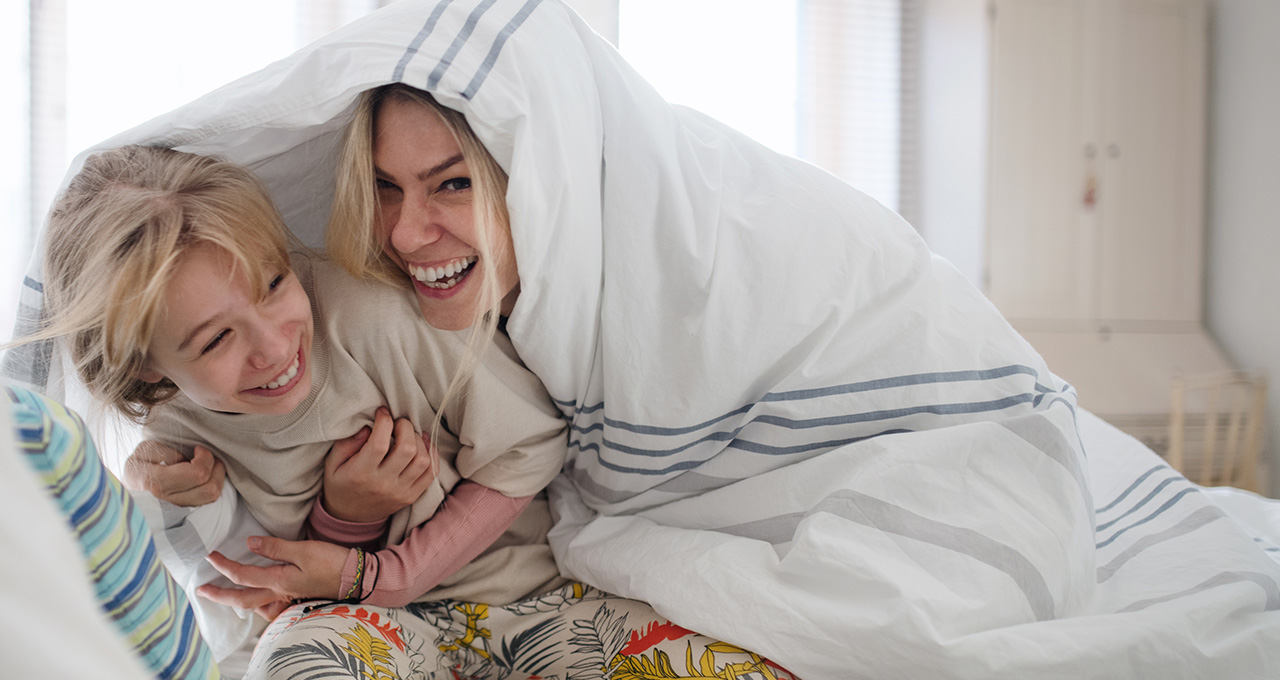Two children are cuddled together under a blanket, creating a cozy atmosphere in a bright room.