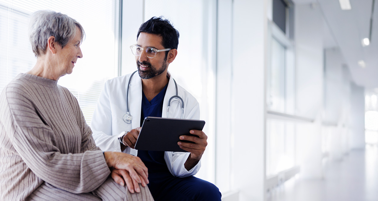 An elderly woman sits with a doctor in a bright medical office, discussing information on a tablet with attentive expressions.