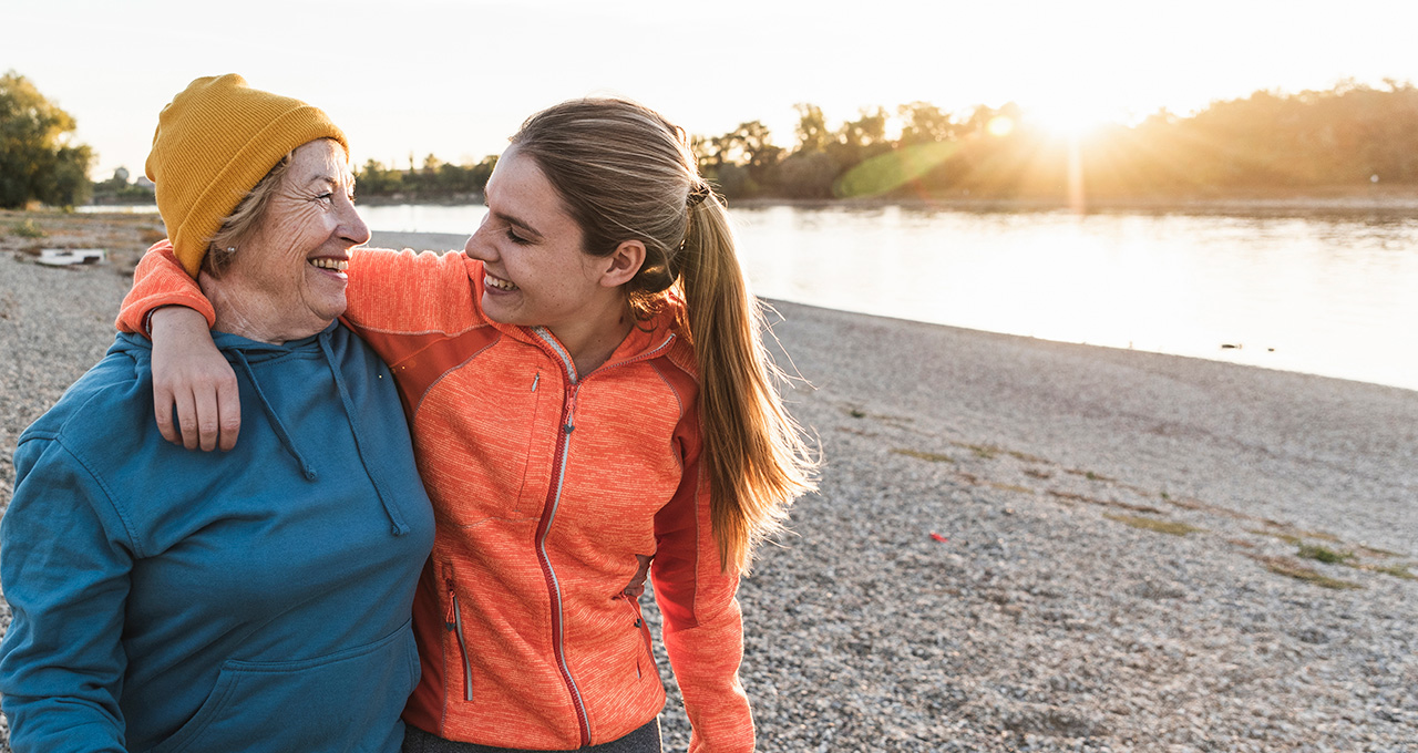 A young woman in an orange jacket embraces an older woman in a blue hoodie, both smiling by the river.