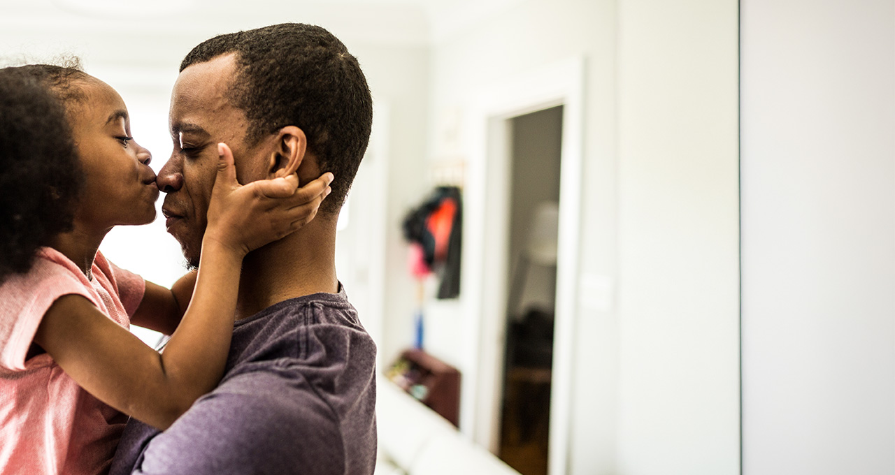 A father holds a child close, both smiling, in a cozy indoor setting with soft lighting.