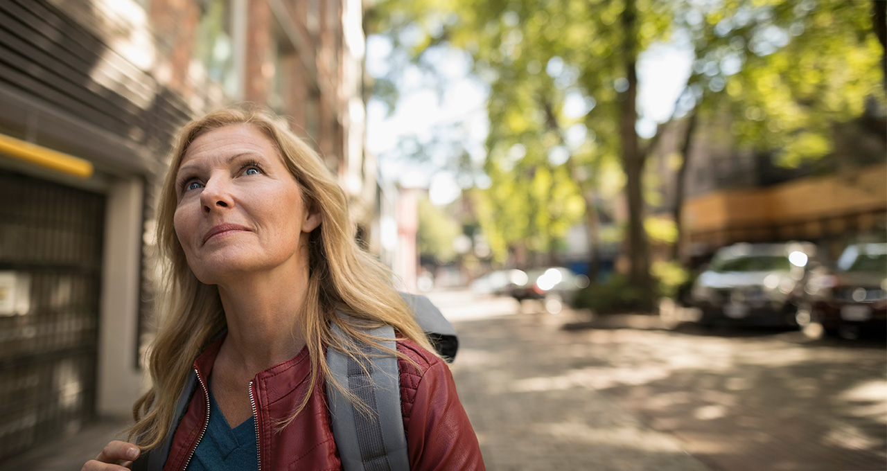 A woman walks down a tree-lined street, wearing a red jacket and carrying a backpack, enjoying the sunny day.